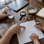 Overhead view of child and adult hands with a pencil, a cup of water, a magnifying glass and beans on a dining table by a window.